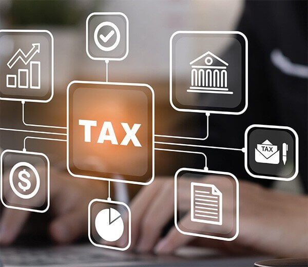 A central square labeled "TAX" surrounded by financial icons like a bar graph, dollar sign, pie chart, and bank, with blurred hands typing on a keyboard in the background.