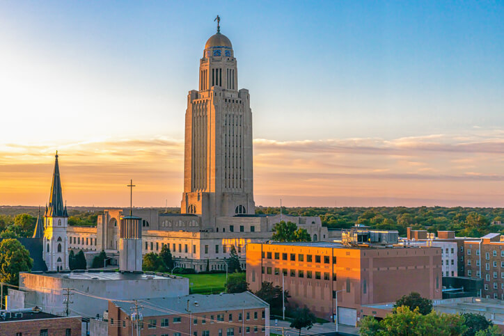 Nebraska State Capitol building in Lincoln at sunset, with its tall tower, dome, and 'The Sower' statue illuminated by warm light. A church steeple and surrounding buildings are visible, with a partly cloudy sky in the background.