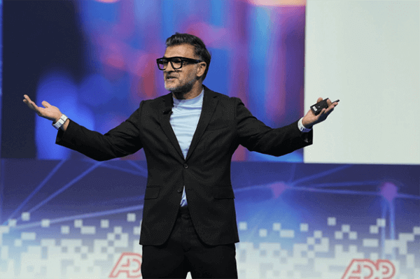 John Sanei stands on a stage with arms outstretched, in front of a digital background with "ADP" visible at the bottom.